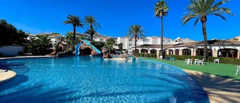 A large swimming pool with a water slide, palm trees, and a building with a tiled roof in the background.