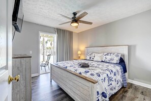 Une chambre à coucher avec un lit, un ventilateur de plafond, un téléviseur fixé au mur et une porte dotée d’une poignée en laiton.