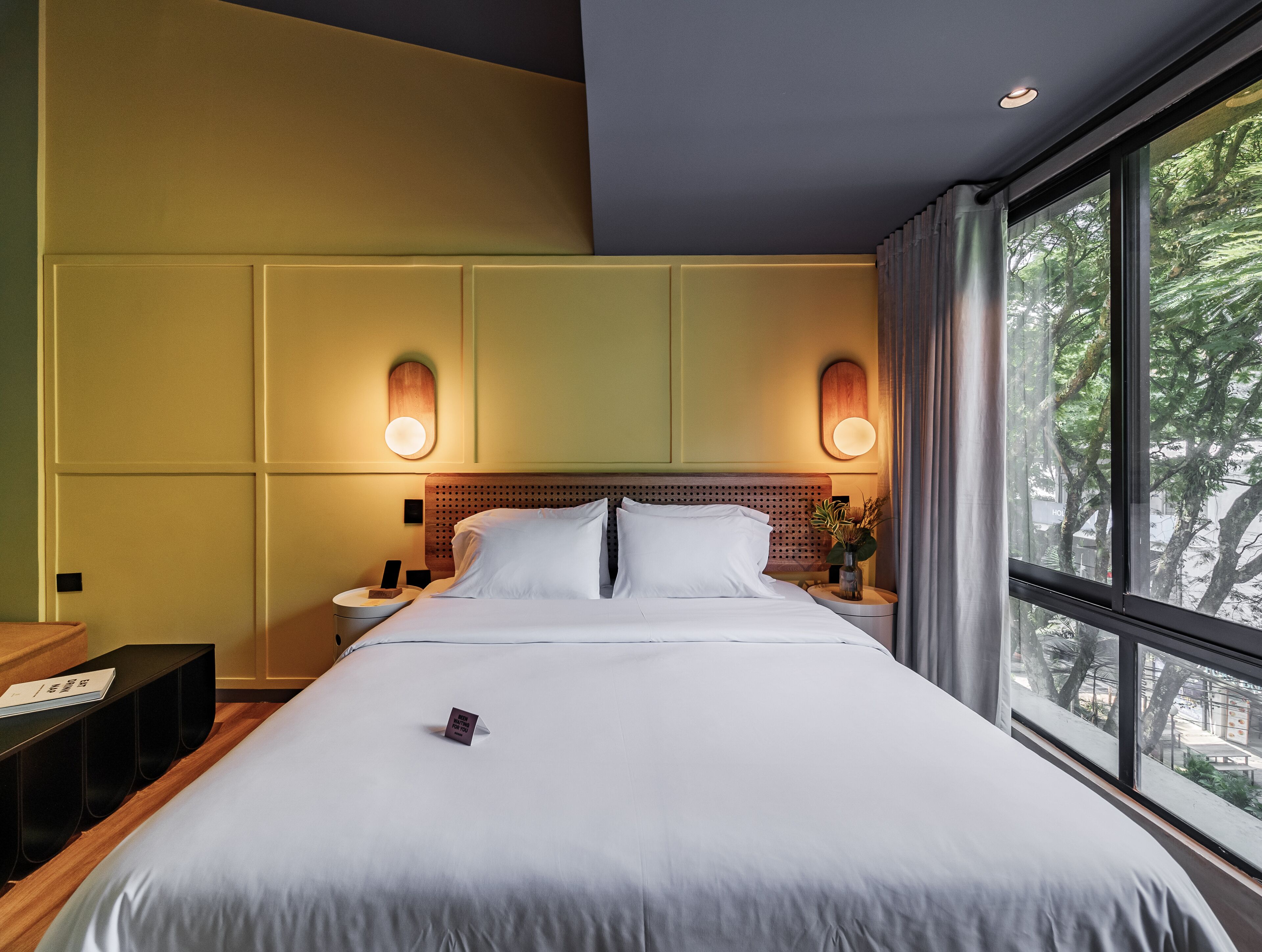 Quarto de hotel com uma cama grande, duas mesas de cabeceira, um criado-mudo, luminária fixada na parede e uma janela com vista para árvores.