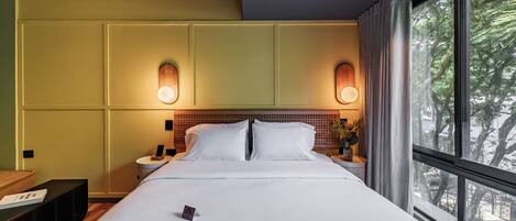 A hotel room with a large bed, two bedside tables, a nightstand, a wall-mounted light fixture, and a window with a view of trees.