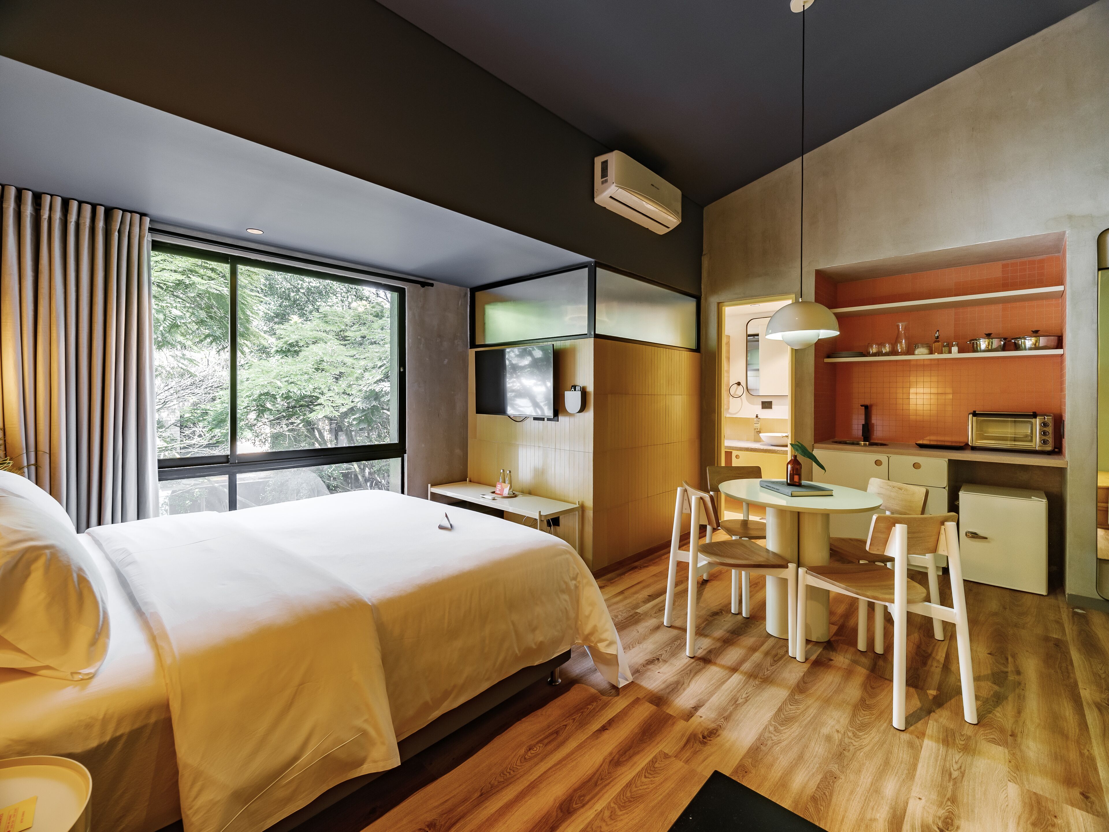 A modern hotel room with a large bed, a desk, a chair, a small kitchenette, and a large window offering a view of trees.