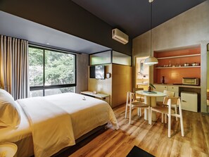 Une chambre dâhĂŽtel moderne dotĂ©e dâun grand lit, dâun bureau, dâune chaise, dâune petite kitchenette et dâune grande fenĂȘtre donnant sur les arbres.