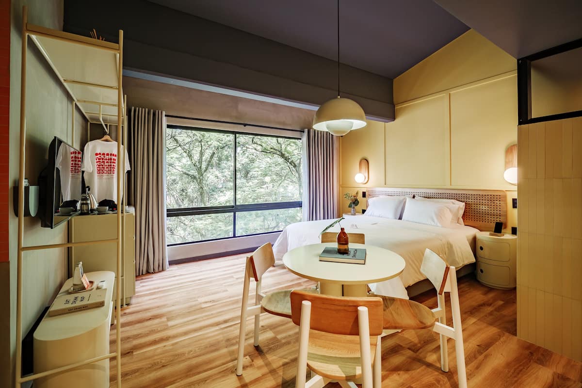 A hotel room with a bed, bedside table, lamp, chair, and a desk with a mirror and a TV.