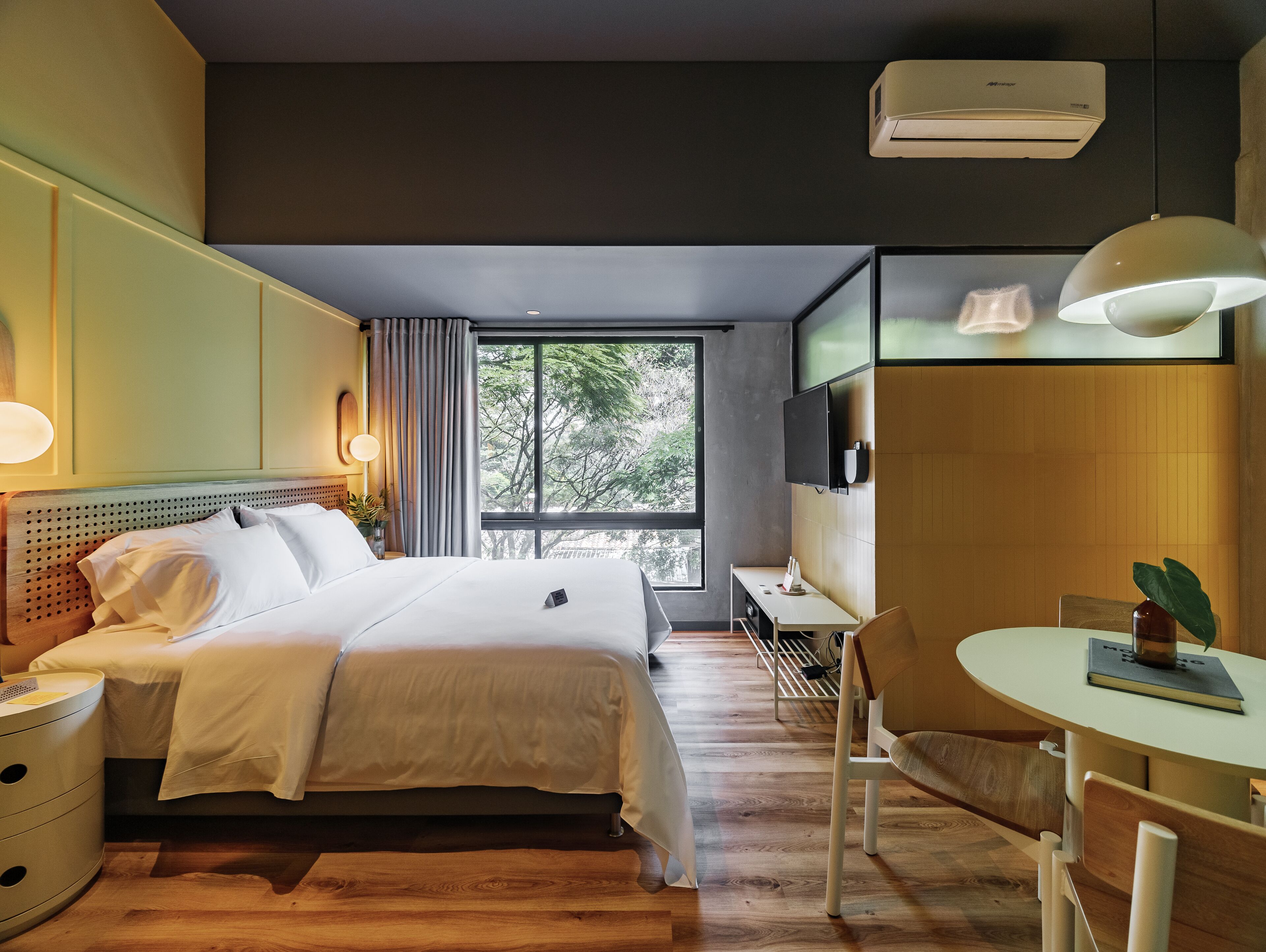 A hotel room with a bed, bedside table, desk, chair, and a large window with a view of trees.