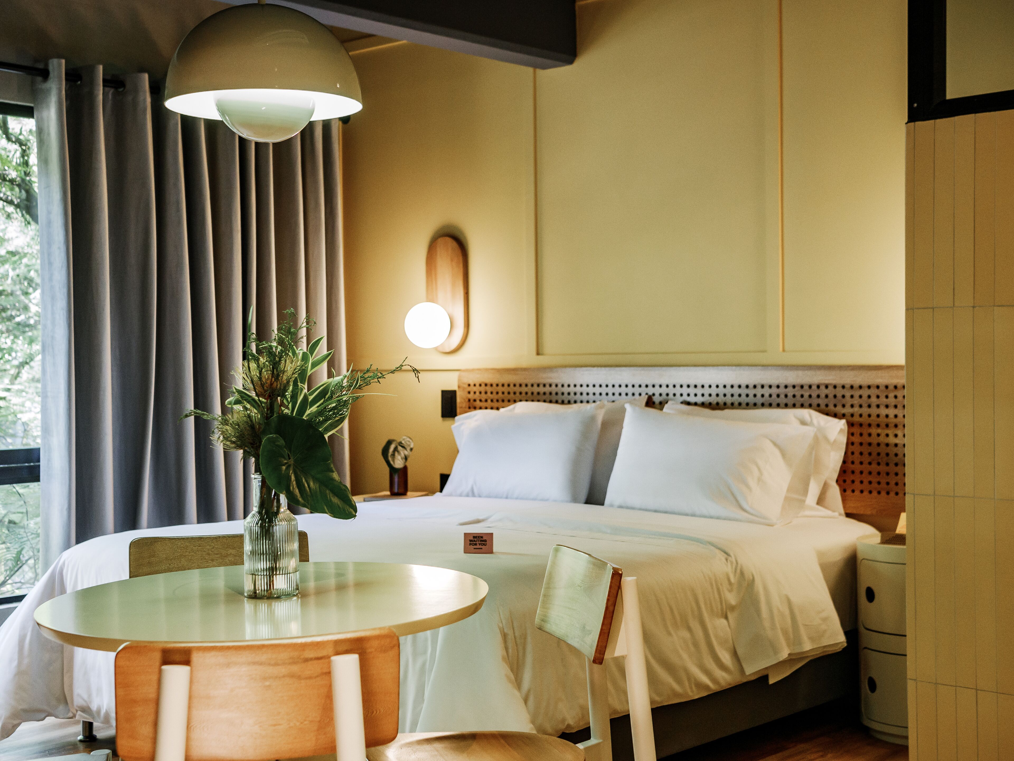 A hotel room with a bed, bedside table, chair, and a round table with a vase of flowers.