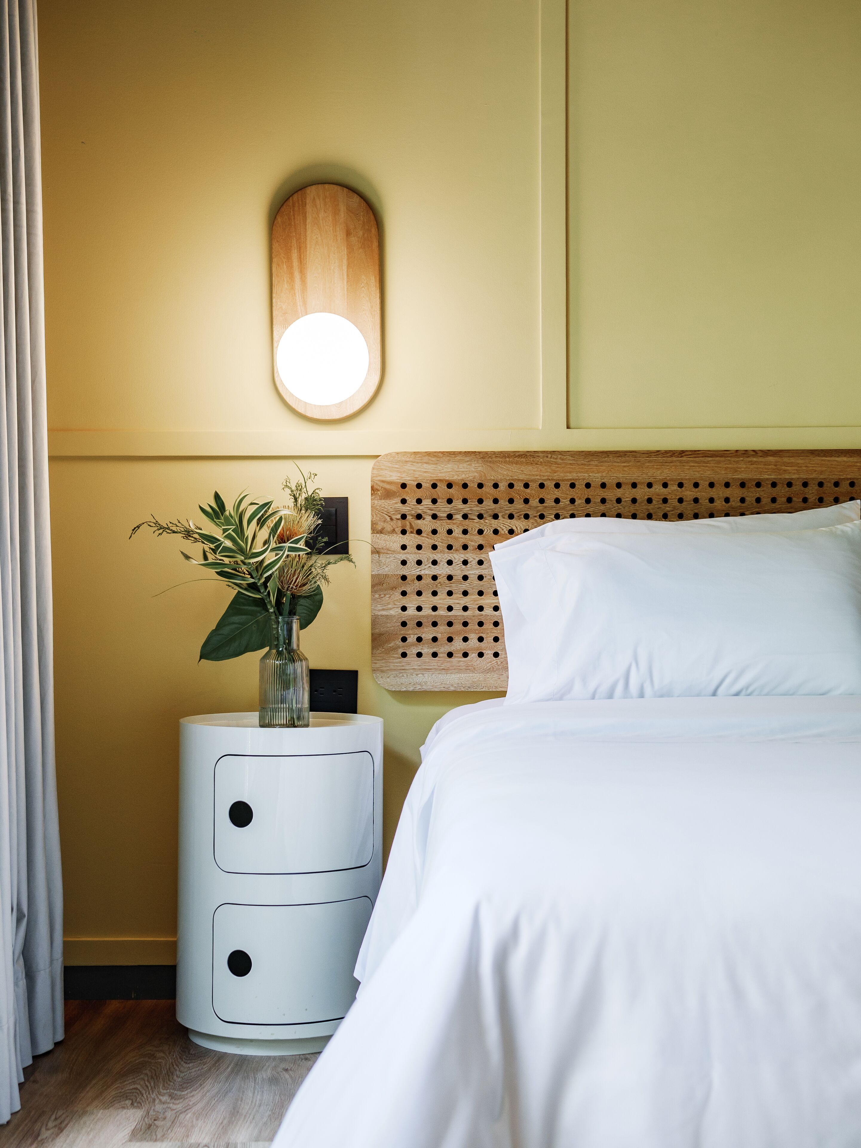 A neatly arranged hotel room with a bed, a bedside table with a vase of flowers, a wall-mounted light, and a wooden headboard.