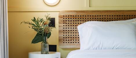 A neatly arranged hotel room with a bed, a bedside table with a vase of flowers, a wall-mounted light, and a wooden headboard.