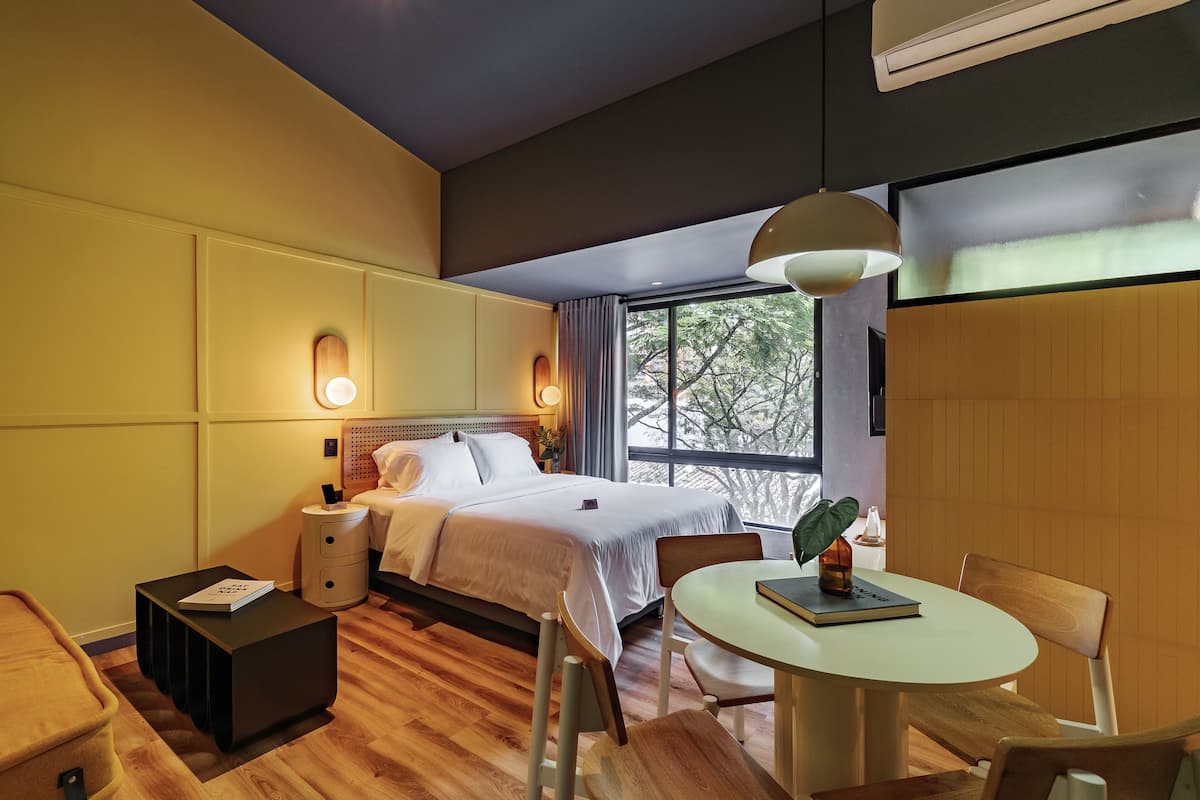 A modern hotel room with a large bed, bedside tables, a round dining table, and a view of trees through a large window.