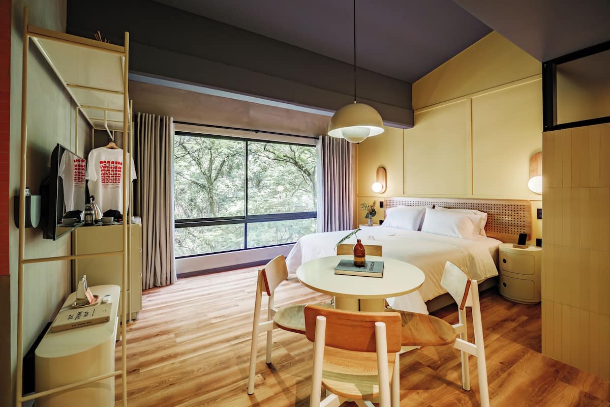 A hotel room with a bed, bedside table, lamp, chair, and a desk with a mirror and a TV.