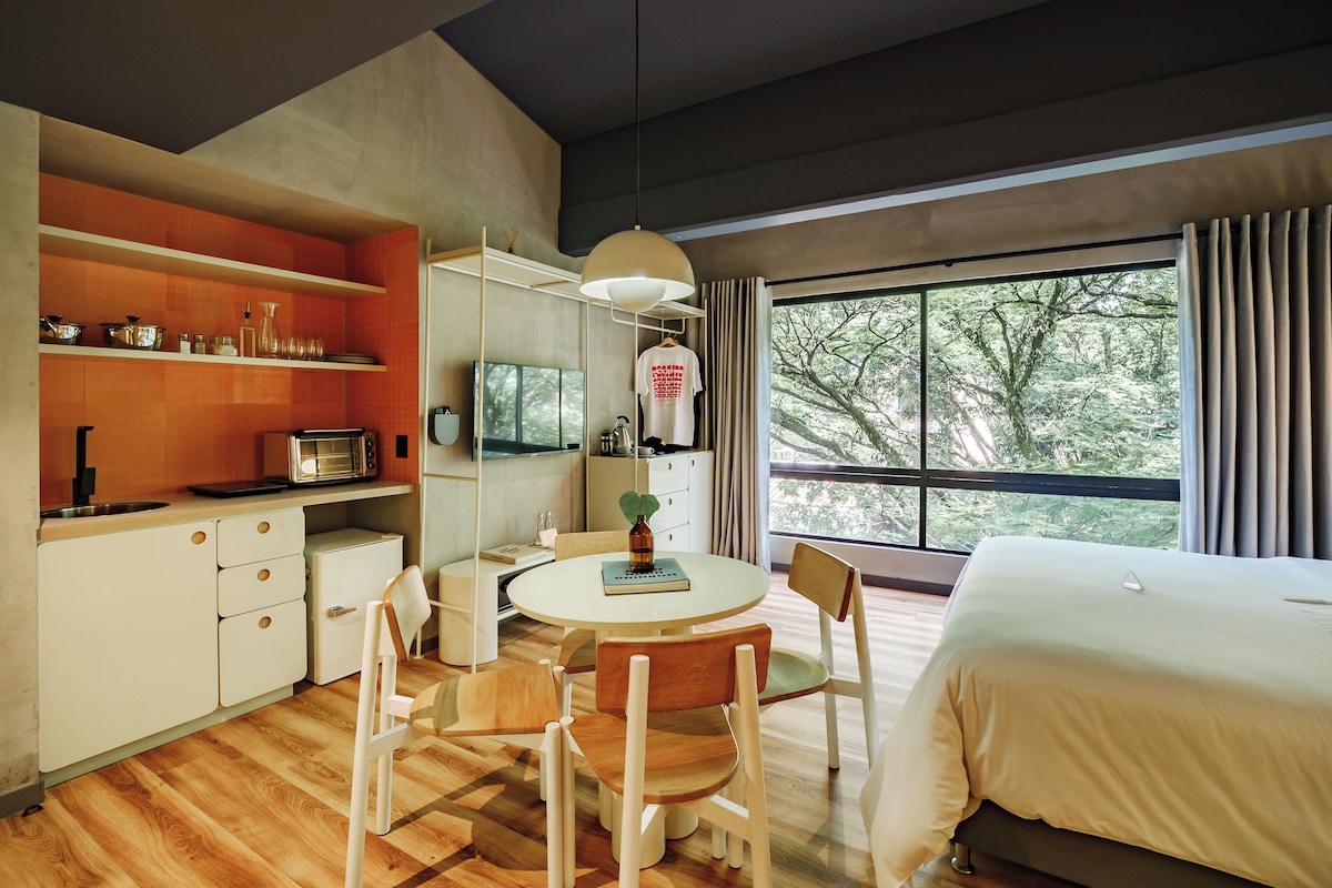 A modern hotel room with a bed, a dining table with chairs, a kitchen area with a stove, and a large window with a view of trees.