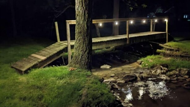 A wooden bridge over a small stream at night, illuminated by string lights. - Your summer getaway- with a hot tub!

 (Millmont)
