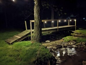A wooden bridge over a small stream at night, illuminated by string lights. - Your summer getaway- with a hot tub!

 (Millmont)