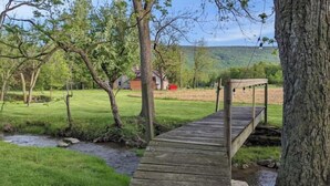 A wooden bridge over a stream in a grassy area with trees and a house in the background. - Your summer getaway- with a hot tub!

 (Millmont)