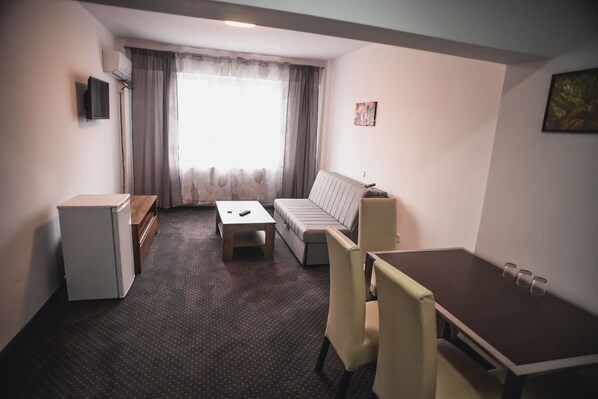 A compact hotel room with a sofa, a small table, a desk, and a refrigerator. - Petrosani Camere de Inchiriat - Hotel Petroshani srl (Petrosani)