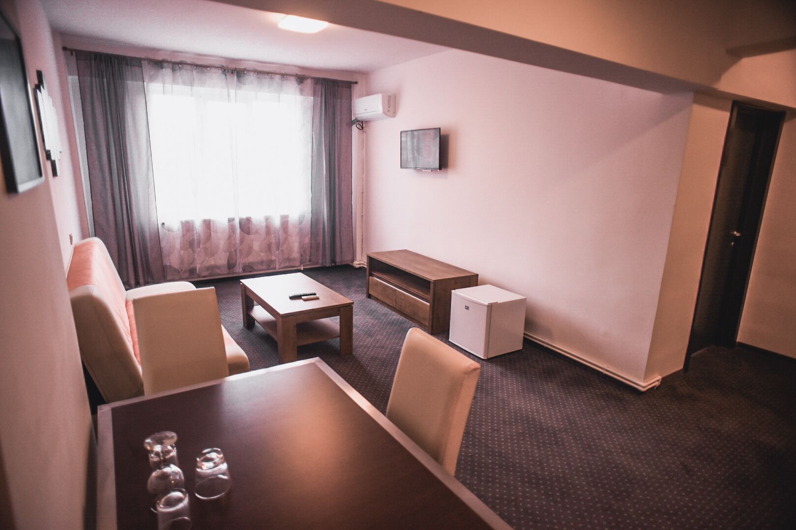 Una habitación de hotel compacta con sofá, una pequeña mesa de comedor, una mesita de centro, televisor y un refrigerador pequeño.