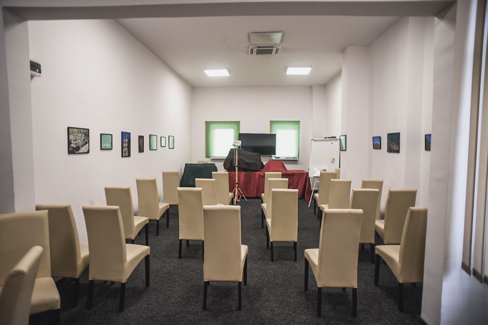 A conference room with beige chairs arranged in rows, a whiteboard, and a projector screen.