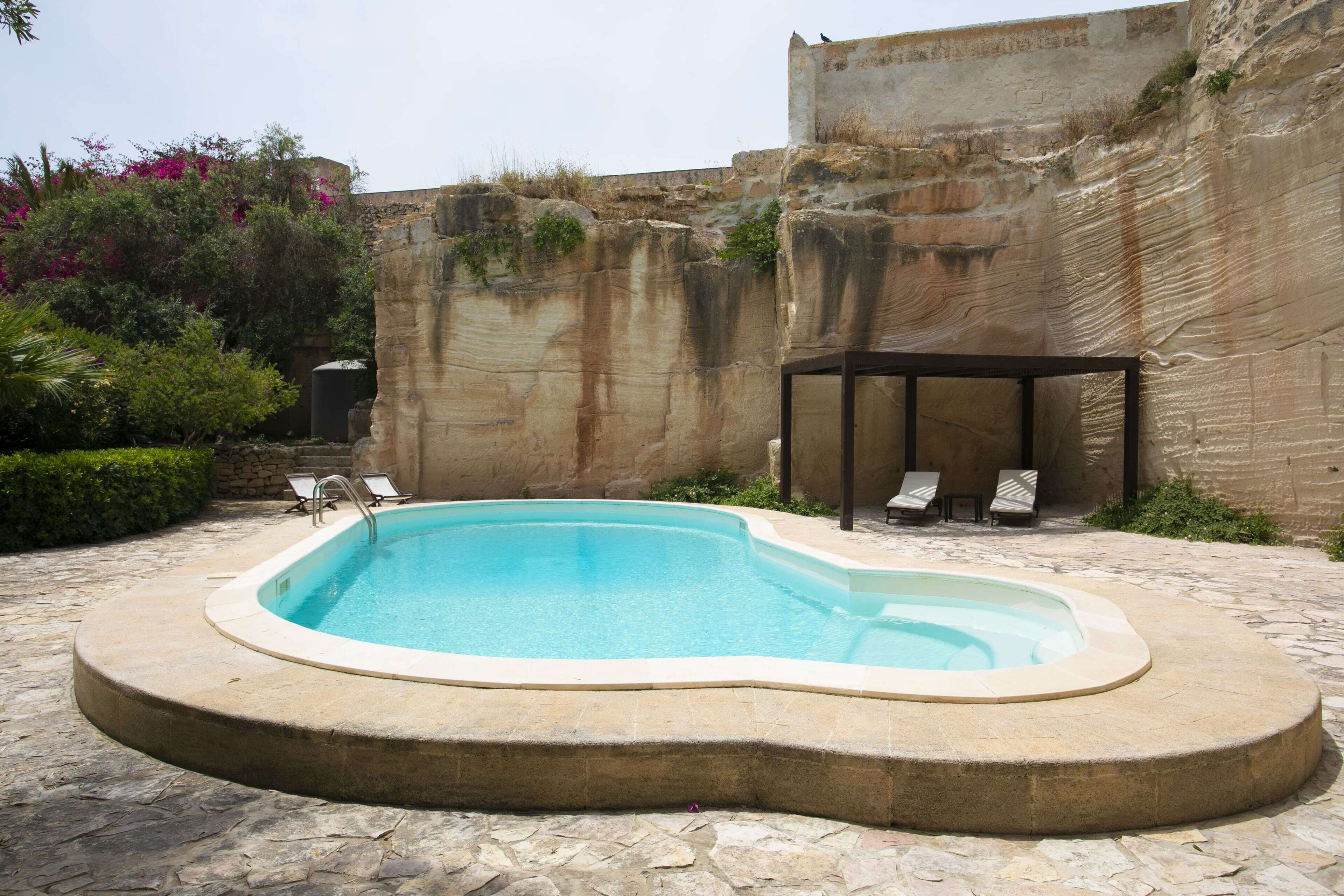 Una piscina con solárium de piedra, rodeada por un muro de piedra y una zona de estar cubierta con sillones de relax.