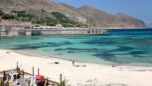 Une plage côtière aux eaux turquoise et limpides, bordée d’un rivage sablonneux, avec une chaîne de montagnes en arrière-plan.