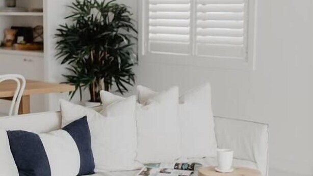 A minimalist white bedroom with a bed, a side table, a potted plant, and a window with shutters. - The Palm House Gerroa (Gerroa)
