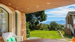 Eine Terrasse mit Tisch und Stühlen, eine Steinmauer und Blick auf eine Wiese mit Bäumen und Bergen.