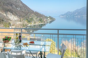 Varanda com mesa posta para refeições, vista para um lago com barcos e paisagem montanhosa.