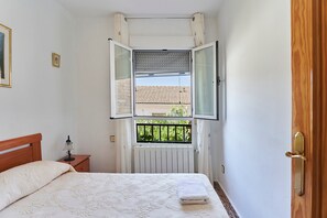 Un dormitorio con cama, mesita de noche, ventana con cortinas y vista a un edificio.