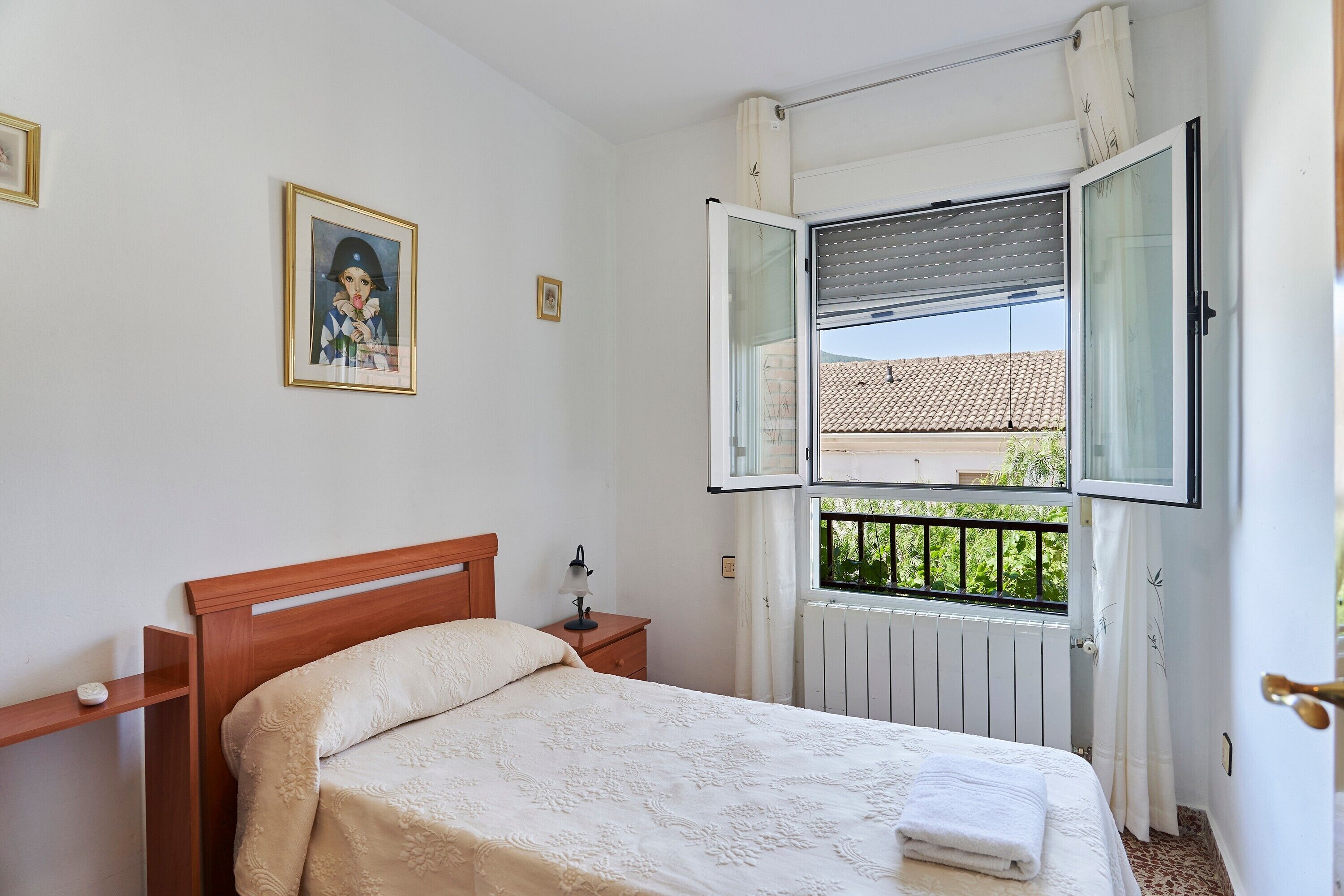 Ein Schlafzimmer mit einem hölzernen Bett, einem Nachttisch, einem Fenster mit geöffneter Jalousie und einem gerahmten Bild an der Wand.