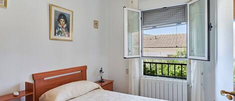 Ein Schlafzimmer mit einem hölzernen Bett, einem Nachttisch, einem Fenster mit geöffneter Jalousie und einem gerahmten Bild an der Wand.