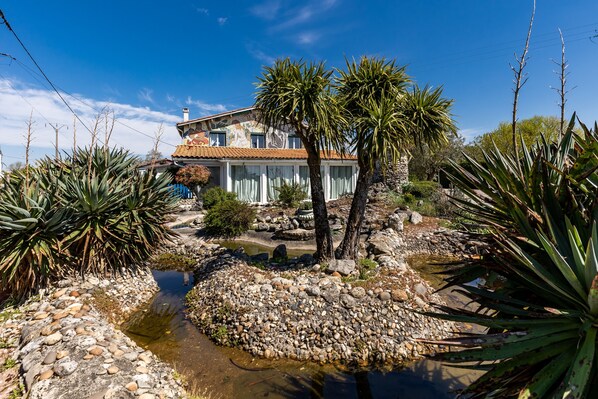 A house with a stone facade, a tiled roof, and a garden with a pond and tropical plants.