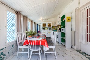 A dining area with a table set for two, white chairs, a potted plant, and a red tablecloth.