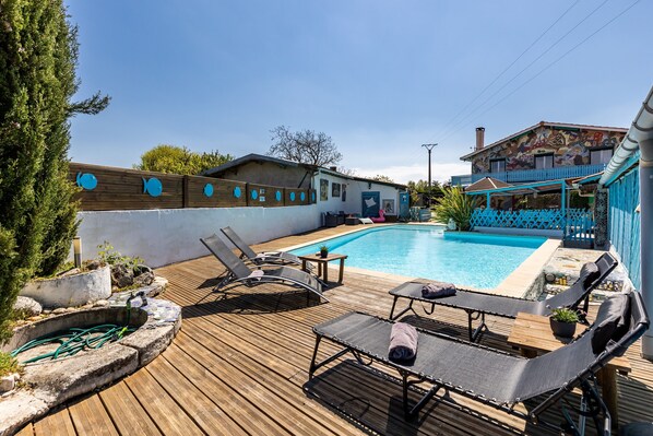 A wooden deck with lounge chairs, a swimming pool, and a house with a colorful mural. - Holiday Home "Gîte 2 à la Maison Mosaïc" with Shared Pool & Wi-Fi (Gaillan en Medoc)