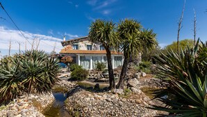 A house with a stone facade, a tiled roof, and a garden with a pond and tropical plants.