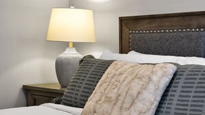 Una cama bien hecha con una colcha blanca y varios almohadones, incluyendo uno de color beige y mullido. Una lámpara con base texturizada se encuentra sobre una mesita de noche junto a la cama.