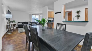 Una sala de estar moderna con una mesa de comedor de madera oscura, sillas negras y una planta grande en maceta. Se observa un sofá y un televisor al fondo.