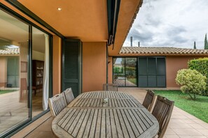 Une terrasse extérieure avec une table et des chaises en bois, une porte vitrée donnant sur un espace abrité, et une vue sur une maison avec un toit en tuiles.