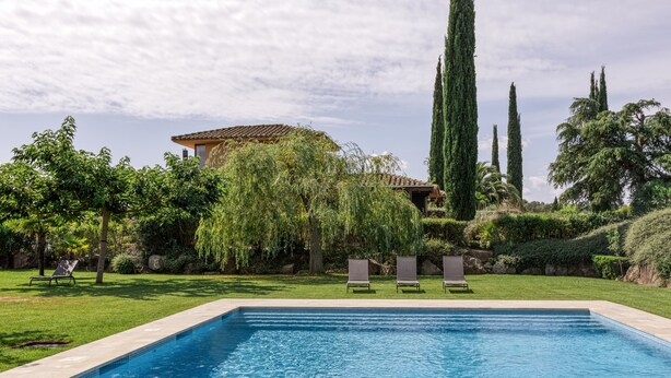 A swimming pool with lounge chairs, a building with a tiled roof, and tall trees in the background. - Holiday Home 'Josi' with Shared Pool, Private Terrace and Wi-Fi (Navata)