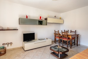 A living room with a TV, a dining table with chairs, a coffee table, and a shelf with decorative items.