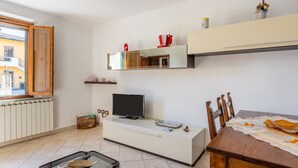 A modern living room with a TV on a white cabinet, a dining table with chairs, a window with a view of buildings, and a shelf with decorative items. - Holiday Home 'Enza's Petite Maison' with Wi-Fi (Staffoli)
