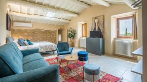 A bedroom with a bed, a blue armchair, a small round table, a radiator, and a window with a view. - Brotes: Chic Apartment in Tuscany, Shared Pool, Hot Tub & Sauna, Wi-Fi, Air Conditioning (Sovicille)