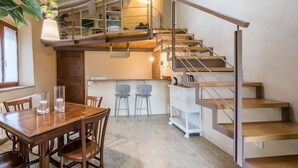 A modern kitchen with a wooden table, two chairs, a staircase with metal railings, and a dining area with a window. - Brotes: Chic Apartment in Tuscany, Shared Pool, Hot Tub & Sauna, Wi-Fi, Air Conditioning (Sovicille)
