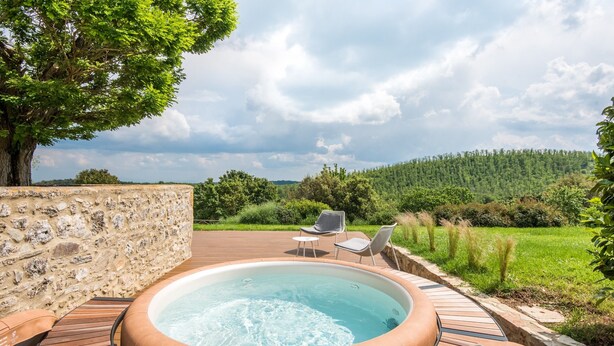 A hot tub with wooden deck seating, surrounded by a stone wall and a view of a forested hillside.