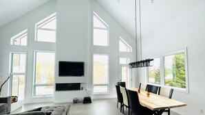 Ein großzügiger Wohnraum mit hohen Decken, großen Fenstern, einem Flachbildfernseher, einem Essbereich mit Holztisch und -stühlen sowie einem grauen Sofa.
