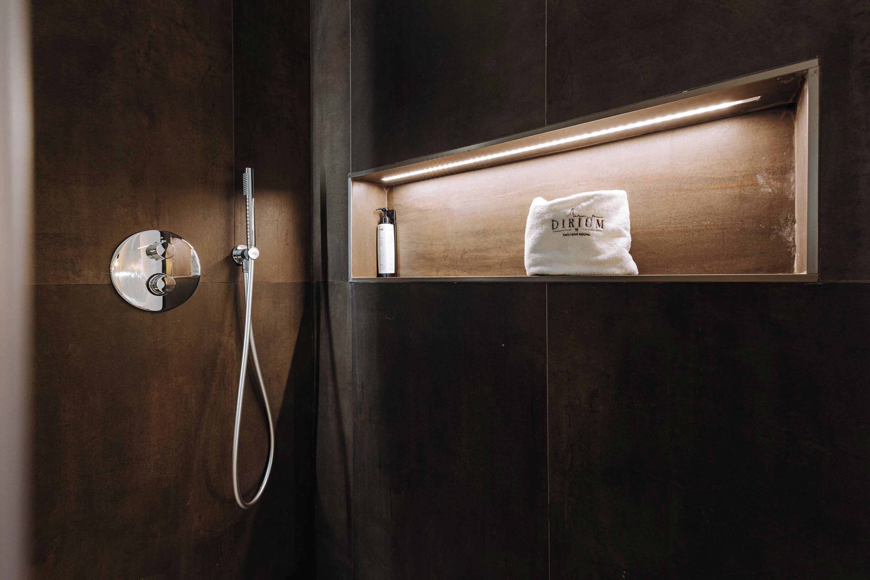 A modern bathroom with a dark wood finish, a built-in shower with a handheld showerhead, and a shelf with a towel and a bottle.