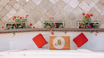 A terrace with a stone wall, red cushions, a wooden table with a placemat, and potted plants with red flowers.