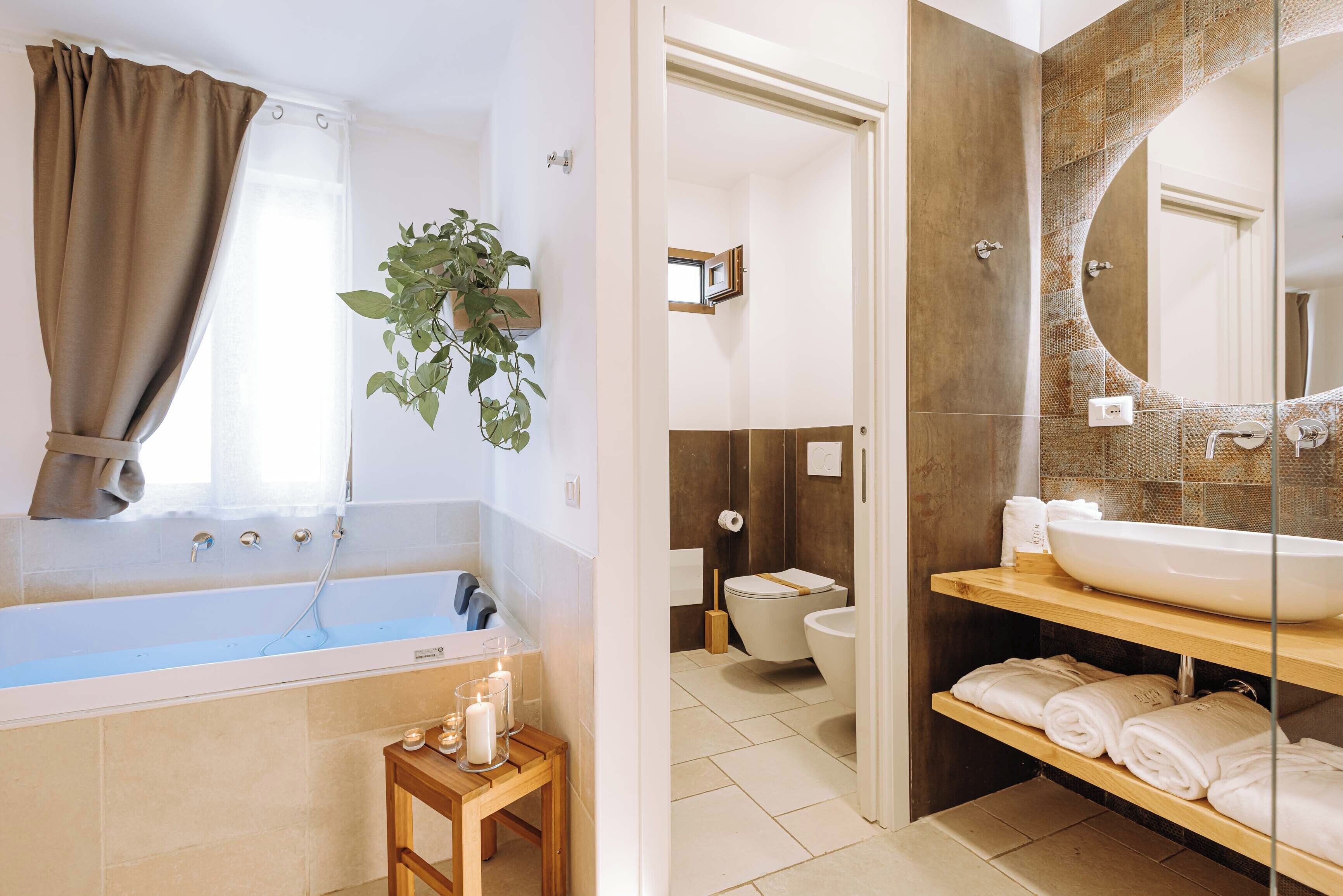 A bathroom with a bathtub, a toilet, and a large mirror. There are towels and a plant on the shelves.