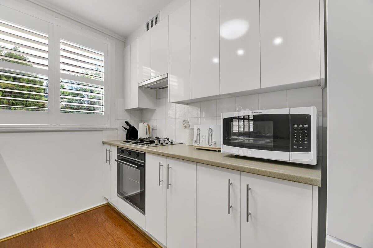 Een moderne keuken met witte kasten, een ingebouwde oven, een magnetron en een raam met zonwering.