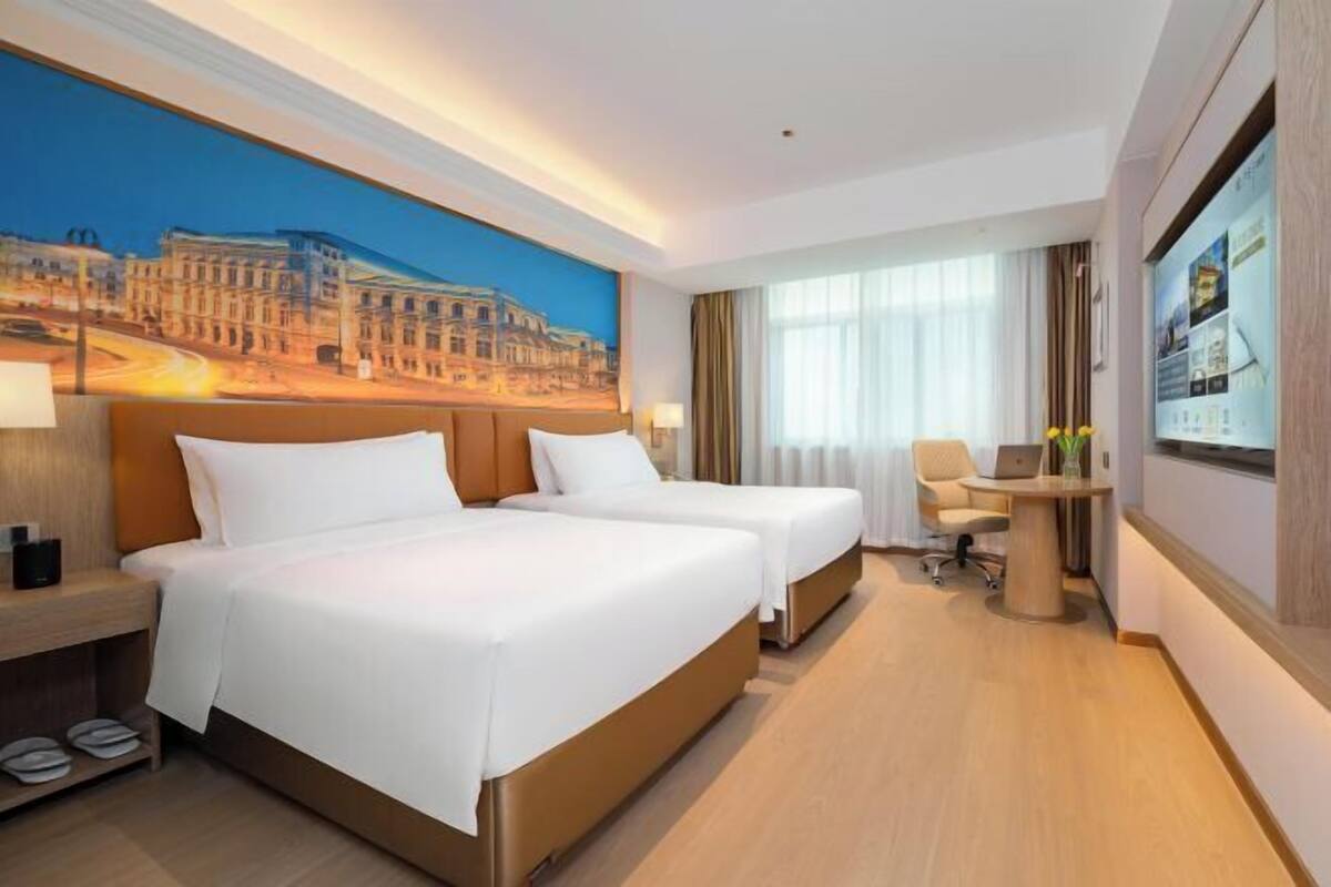 A hotel room with two beds, a desk with a laptop, a chair, and a large window with curtains.