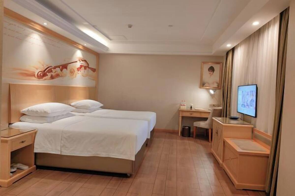 A hotel room with a double bed, a desk with a chair, a television, and a small trash bin.