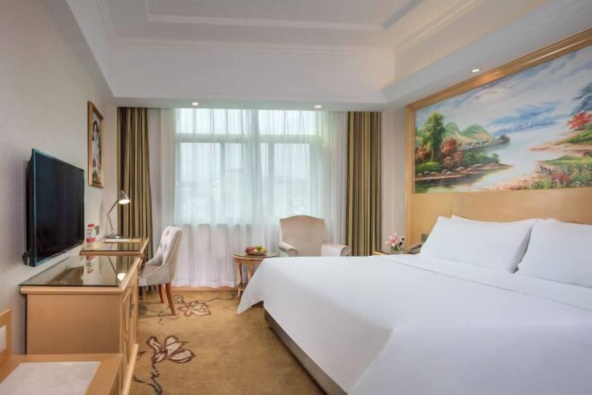 A hotel room with a large bed, a desk with a chair, a television, and a painting on the wall.