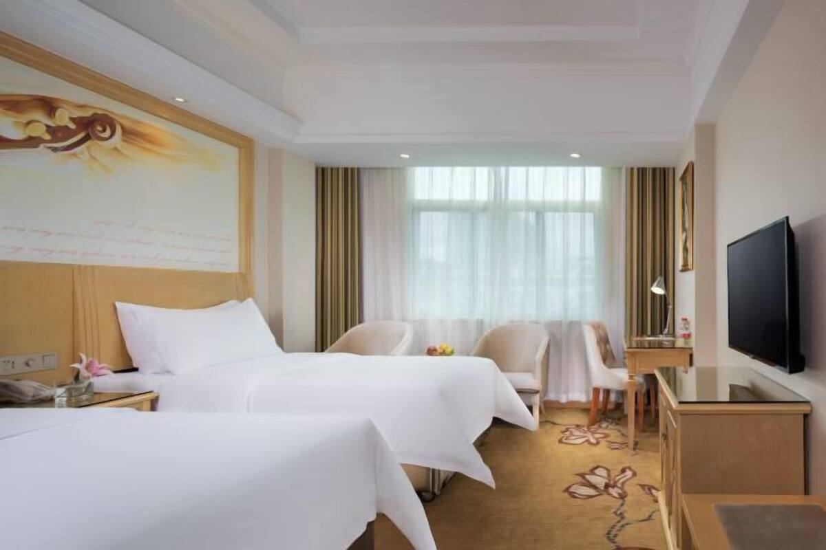 A hotel room with two beds, a desk, a TV, and a large window with curtains.